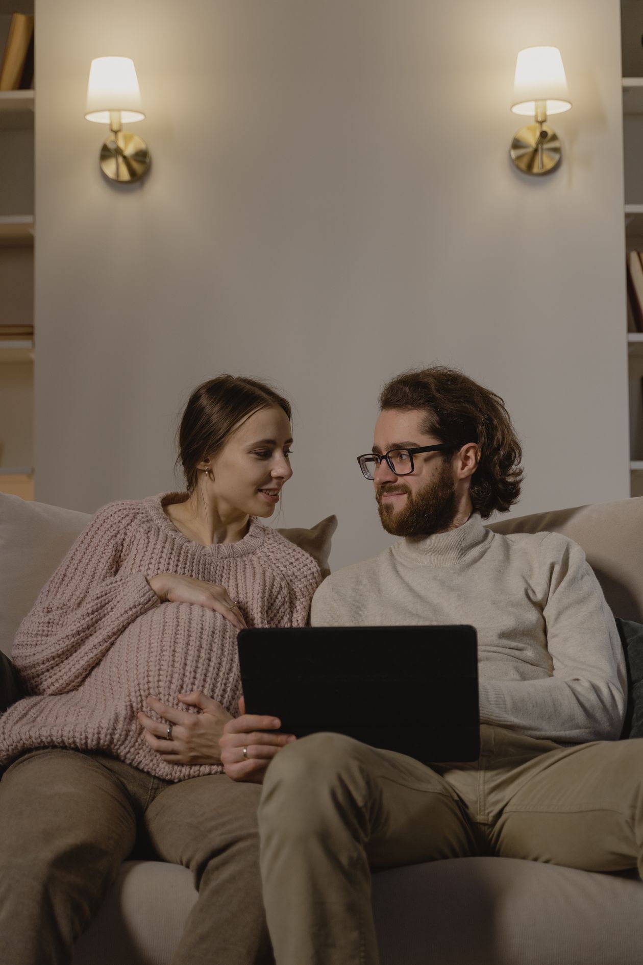A young expecting couple sitting on a sofa, looking at each other.