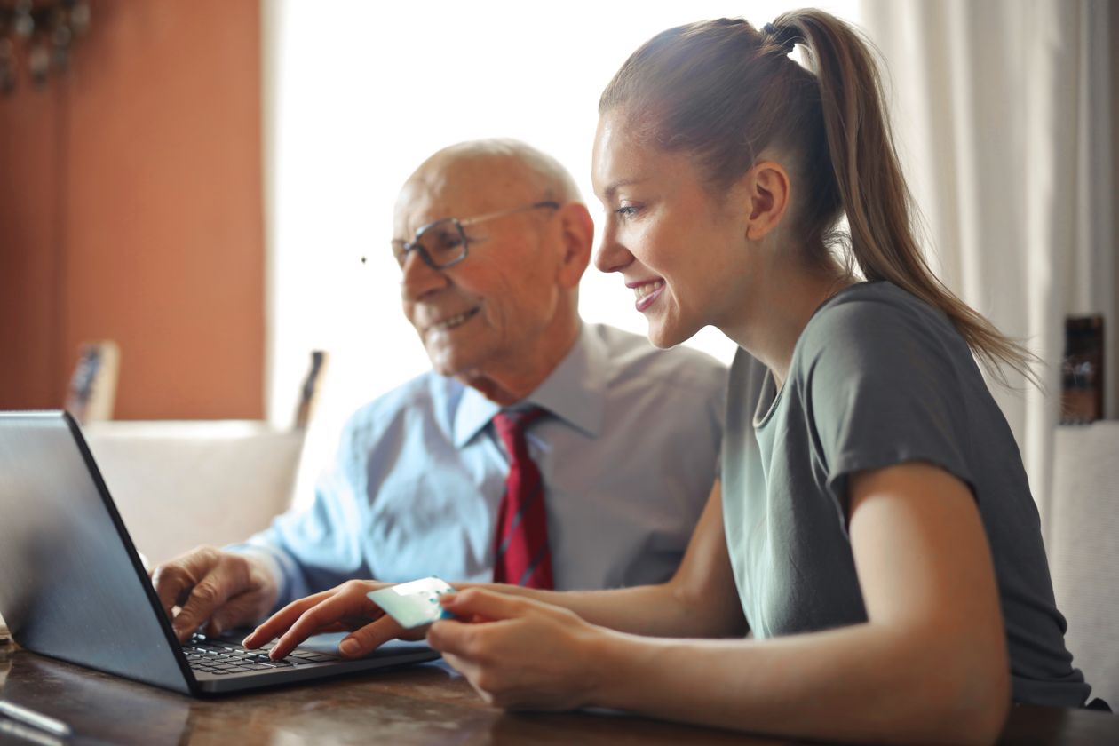 A younger woman helping an older man with paperwork on a laptop at home.